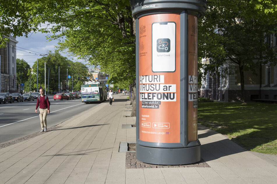 Ein Werbeplakat für die lettische Corona-Warn-App hängt an einer nahezu menschenleeren Straße in einer Litfaßsäule.