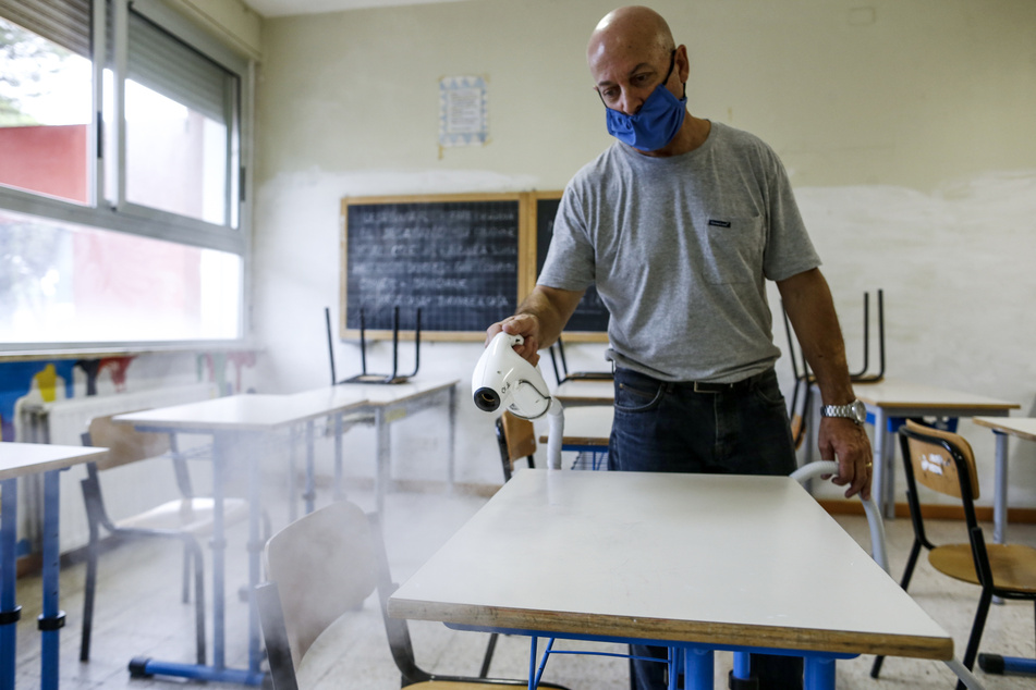 Ein Mann mit Mundschutz desinfiziert Schreibtische in einem Klassenzimmer.