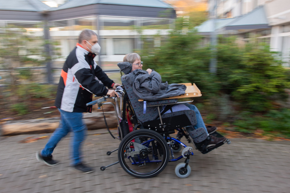 Ein Mitarbeiter der Assistenz schiebt einen Bewohner in seinem Rollstuhl über einen Hof an einer Diakonie-Wohnstätte.