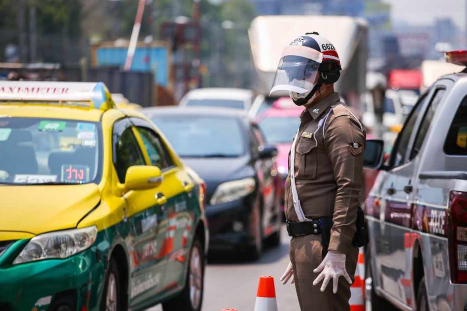 Thailand, Bangkok: Ein Polizeibeamter, der aufgrund der Coronapandemie einen Gesichtsschutz und Schutzhandschuhen trägt, hält an einem Kontrollpunkt Fahrzeuge an.