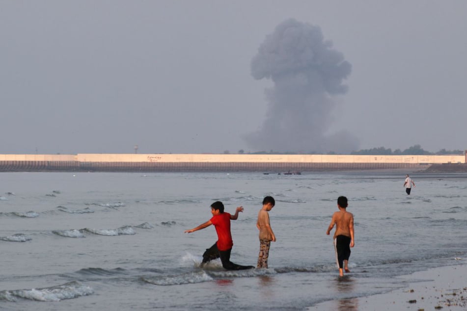 Drei Jungen spielen im seichten Wasser der Straße von Hormus, während im Hintergrund vor der Küste von Bandar Abbas eine Rauchwolke nach einer Explosion aufsteigt.