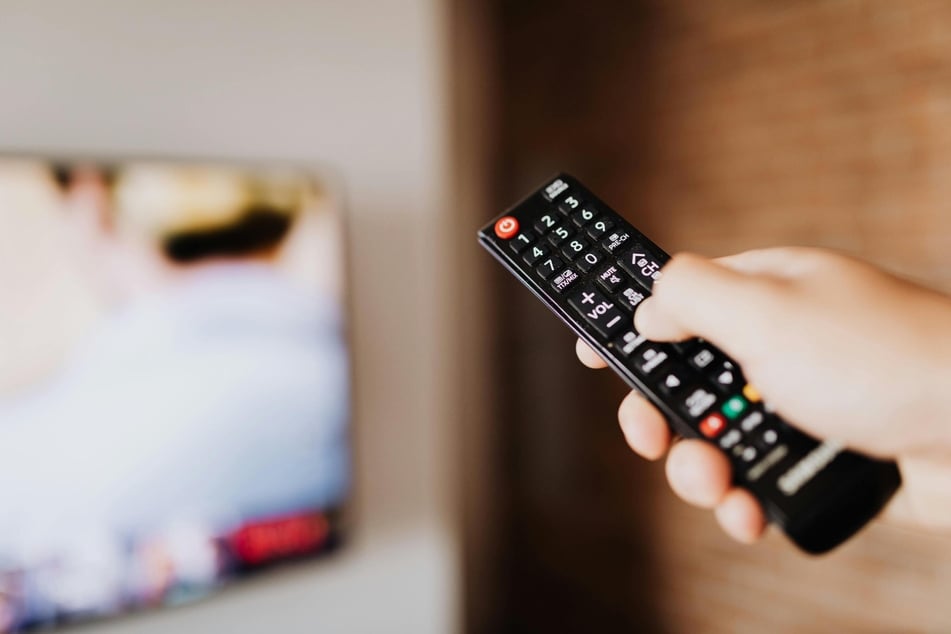 Nahaufnahme einer Hand, die eine schwarze Fernbedienung auf einen unscharfen Fernseher im Hintergrund richtet