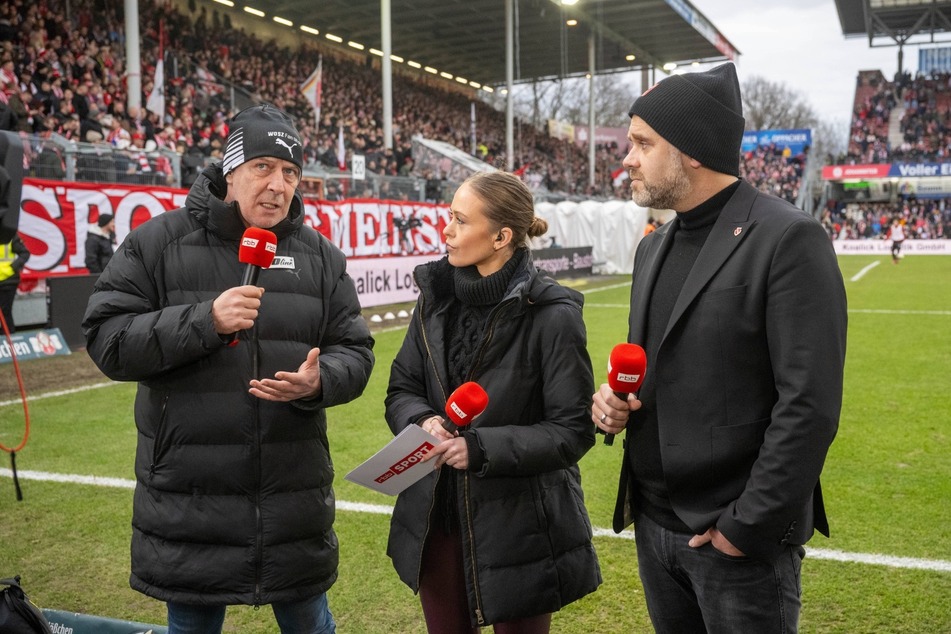 Auch beim Topspiel der 3. Liga ist Mario Basler (l.) ein gefragter Mann im Halbzeit-Interview des rbb. Auf dem Kopf: Eine Mütze des Wosz-Fanshops.