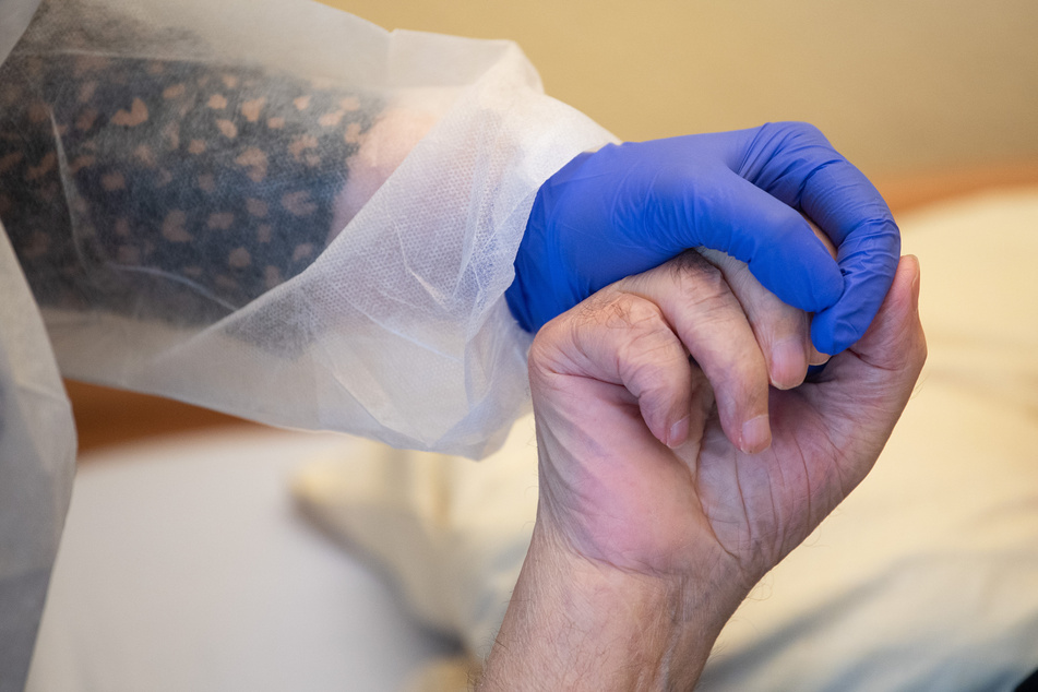 Eine Altenpflegerin in Schutzkleidung hält die Hand eines Bewohners. Die Zahl der Neuinfektionen steigt in Deutschland weiterhin an - auch immer mehr Todesfälle gibt es zu beklagen.