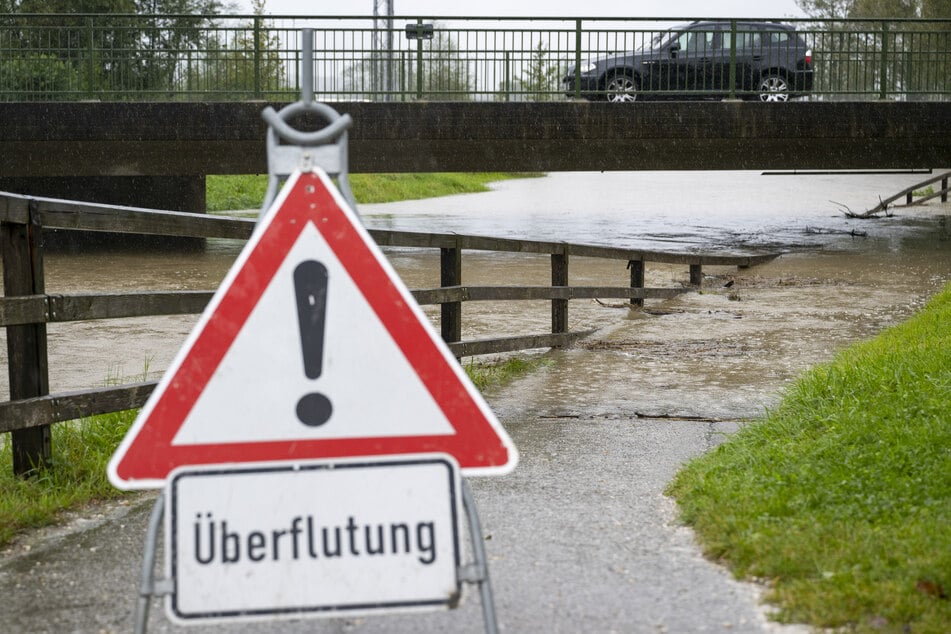 Ein Schild mit der Aufschrift "Überflutung“"steht in Bernau am Chiemsee an einem Weg entlang eines Bachlaufs.