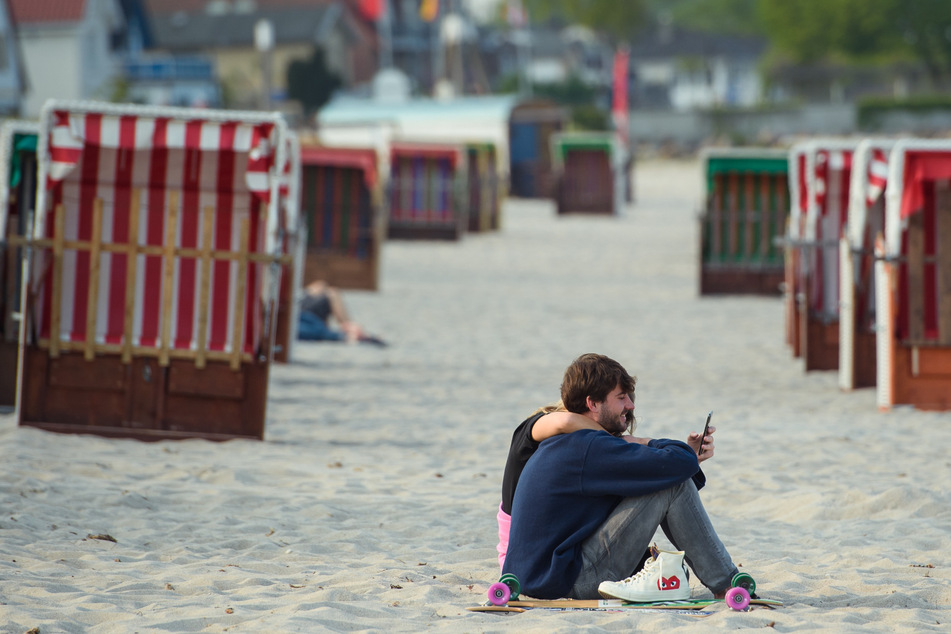 Schleswig-Holstein, Scharbeutz: Ein Paar sitzt im Sand vor zwei Reihen geschlossener Strandkörbe am Strand der Gemeinde Scharbeutz im Kreis Ostholstein.