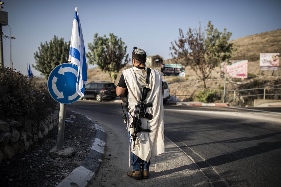 Ein rechtsgerichteter israelischer Siedler trägt eine Waffe am Haupteingang der palästinensischen Stadt Nablus im nördlichen Westjordanland, während rechtsgerichtete israelische Siedler beten.