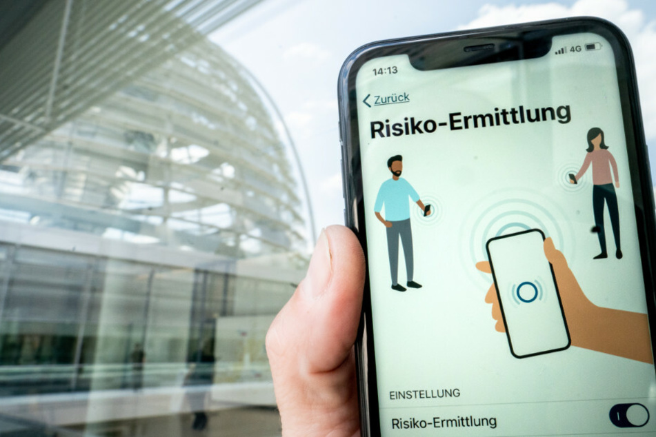 Die Corona-Warn-App mit der Seite zur Risiko-Ermittlung ist im Display eines Smartphone vor der Kuppel des Reichstagsgebäudes zu sehen. Am Montag soll die App um zwei neue Funktionen erweitert werden.