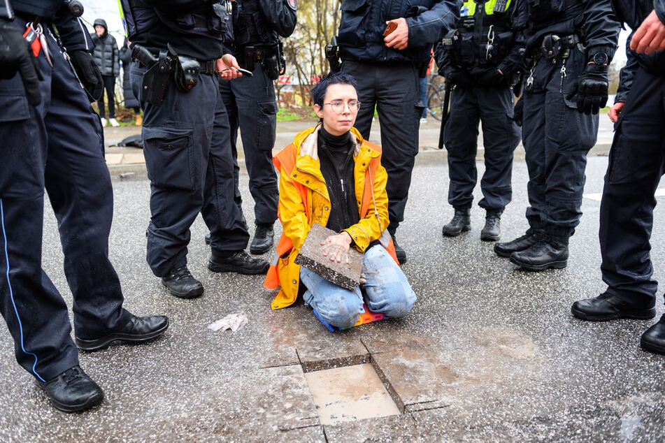 Eine Aktivistin sitzt mit einem Betonteil an ihrer Hand zwischen Polizisten auf der Fahrbahn. Klimaaktivisten der Bewegung «Letzte Generation« haben am 25.03.2023 die Elbbrücken stadteinwärts blockiert.