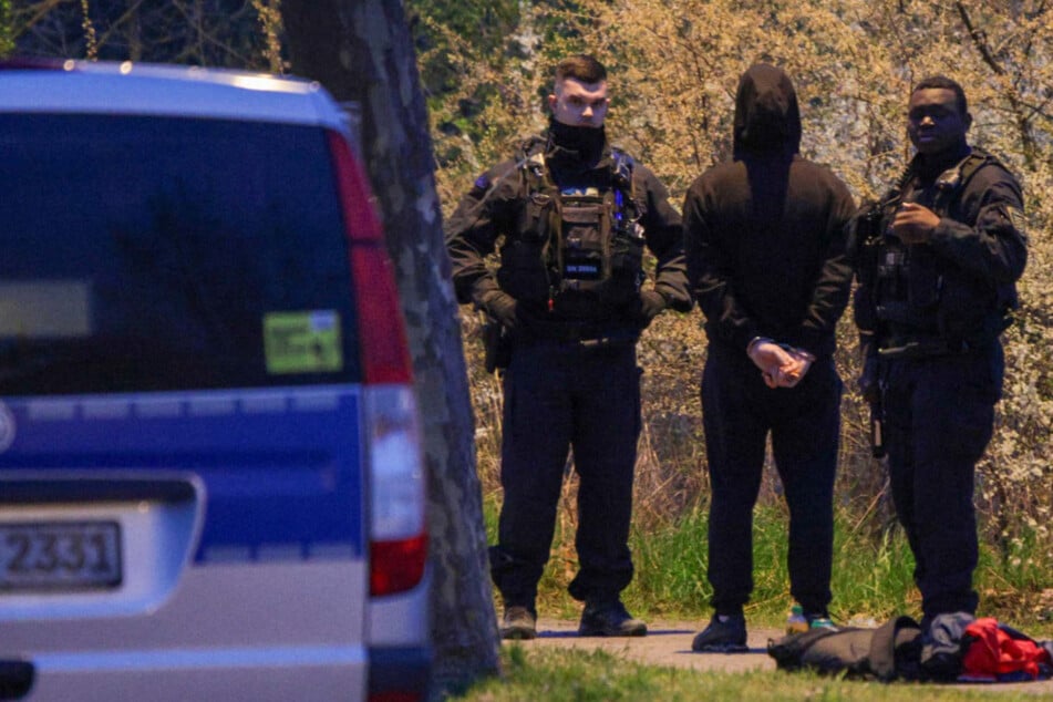 Bilder zeigen unter anderem eine Person in Handschellen, die von mehreren Beamten bewacht wird.