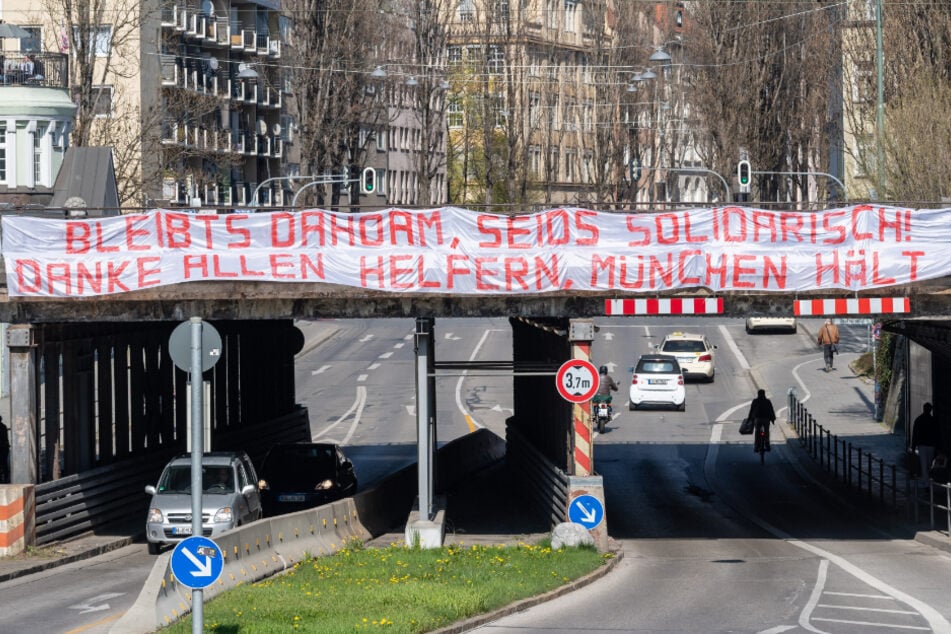 Ein Banner mit der Aufschrift "Bleibts Dahoam, seids solidarisch! Danke allen Helfern, München hält zam!" hängt an einer Eisenbahnbrücke in München.