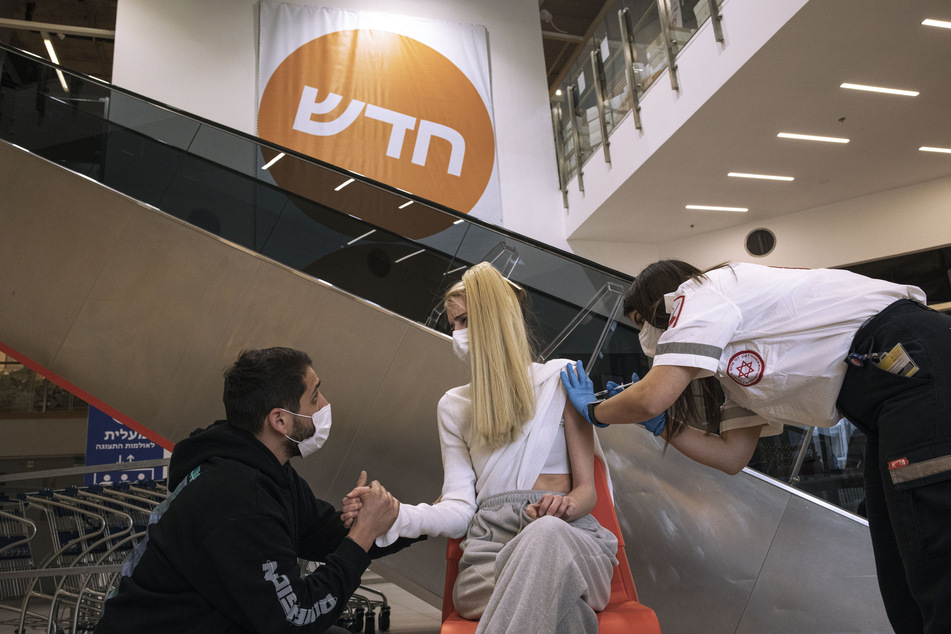 Ein israelischer Sanitäter des medizinischen Dienstes Magen David Adom (r) injiziert einer Frau eine Impfdosis des Corona-Impfstoffs des Mainzer Pharmaunternehmens Biontech und seines US-Partners Pfizer in einer Ikea-Filiale in Rishon Lezion, während ein Mann die Hand der Frau hält.