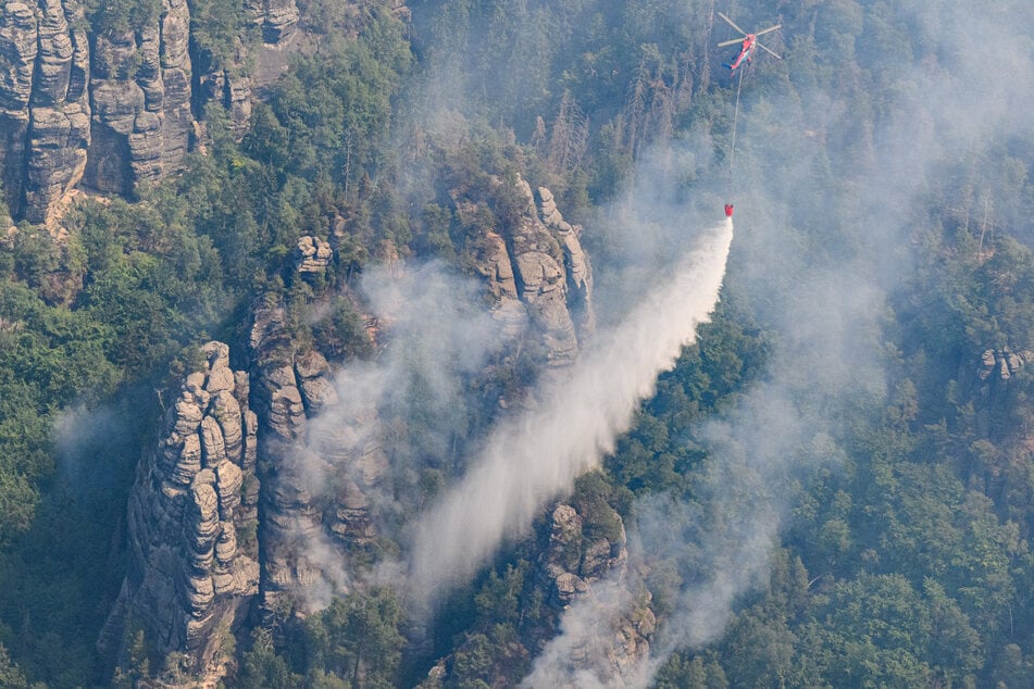 Ein Lastenhubschrauber aus Österreich fliegt mit einem Löschwasser-Außenlastbehälter, um einen Waldbrand in der Sächsischen Schweiz zu löschen.