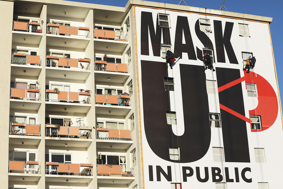 Arbeiter bringen ein Plakat an einer Hauswand an mit der Aufschrift "Mask Up in Public" (deutsch: Masken auf in der Öffentlichkeit).