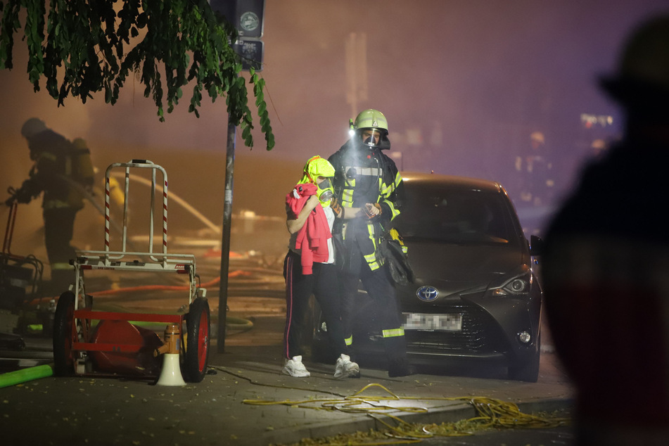 Ein Feuerwehrmann begleitet eine Person, die eine Rettungshaube trägt, von dem brennenden Haus weg.