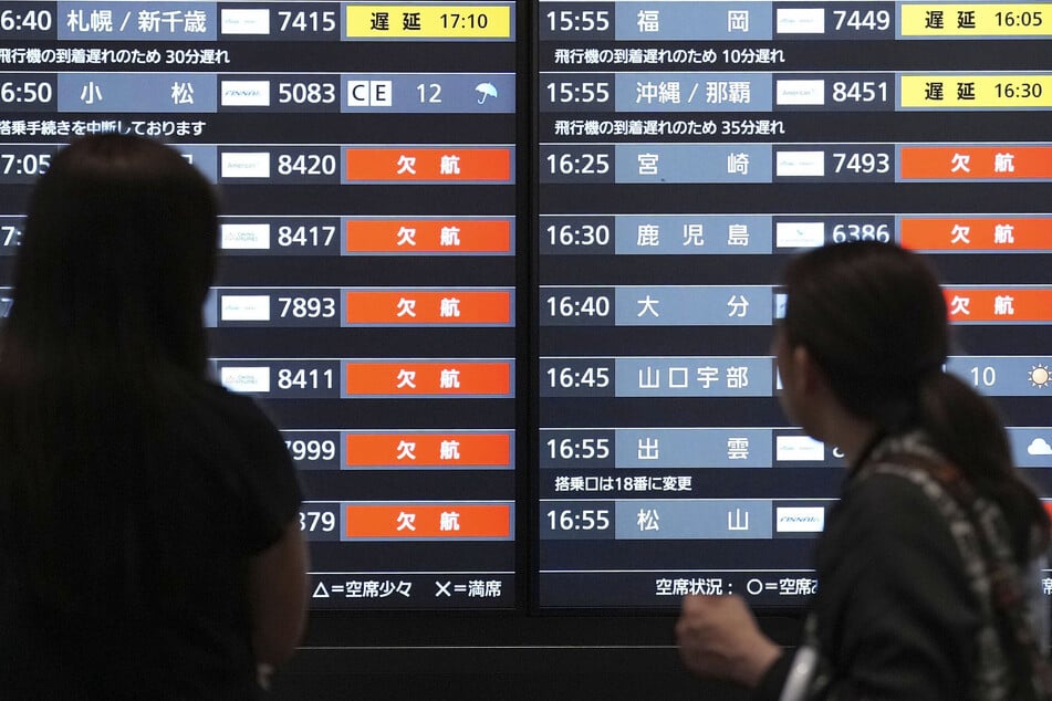 Passagiere am Flughafen Haneda in Tokio blicken auf eine Fluginformationsanzeige, auf der Flugverspätungen und -ausfälle angezeigt werden.