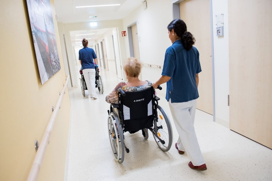 Zwei Pflegerinnen schieben Patienten mit Rollstühlen über einen Krankenhausflur.