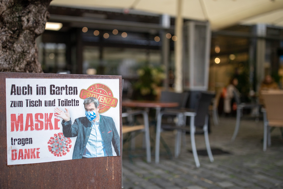 Ein Schild mit der Aufschrift "Auch im Garten, zum Tisch und Toilette Maske tragen - Danke" und dem Konterfei des bayerischen Ministerpräsidenten mit Maske hängt am Eingang zum Außenbereich eines Restaurants.