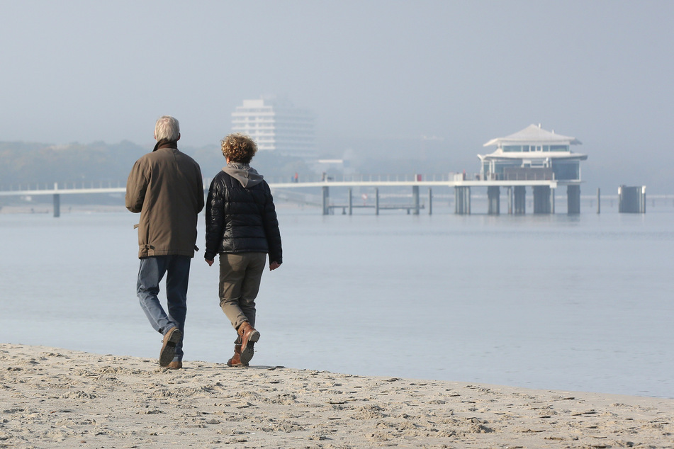 Bei Sonne und leichtem Nebel gehen zwei Spaziergänger am Timmendorfer Strand entlang.