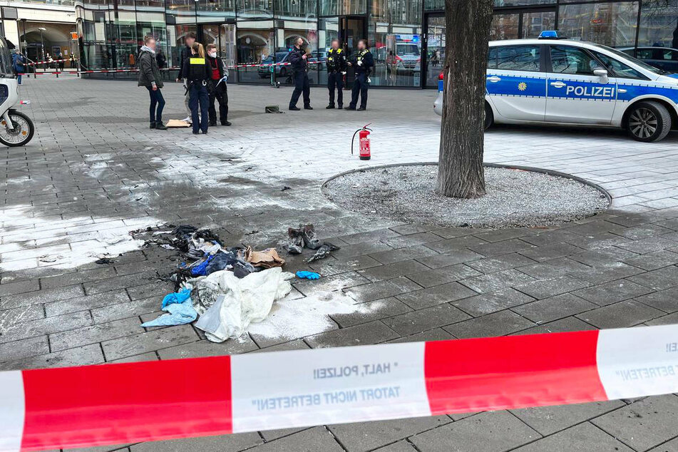 Polizisten stehen am Ort des Geschehens auf dem Berliner Alexanderplatz. Auf dem Boden liegen verkohlte Kleidungsstücke. Ringsherum ist Feuerlöschschaum zu sehen.