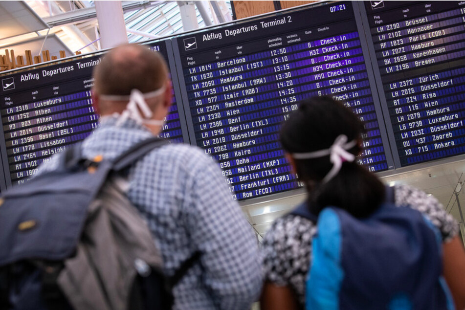 Reisende mit Mundschutz schauen auf eine Anzeigetafel am Münchner Flughafen, auf der zahlreiche internationale Flugziele gelistet sind.