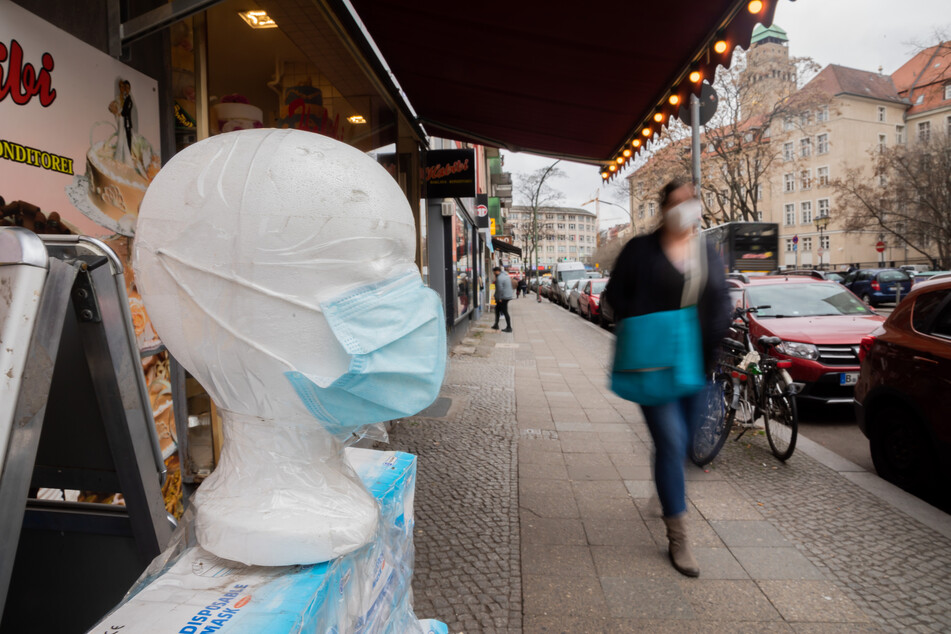 Eine medizinische Maske wird vor einem Geschäft in Berlin-Neukölln ausgestellt, während eine Passantin vorbeigeht.