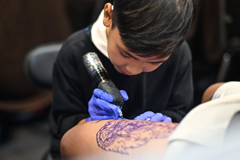 Wielding a cumbersome tattoo gun with his small hands swamped in surgical gloves, Nine-year-old Napat Mitmakorn expertly inks the pattern of a fanged serpent on a man's upper thigh.