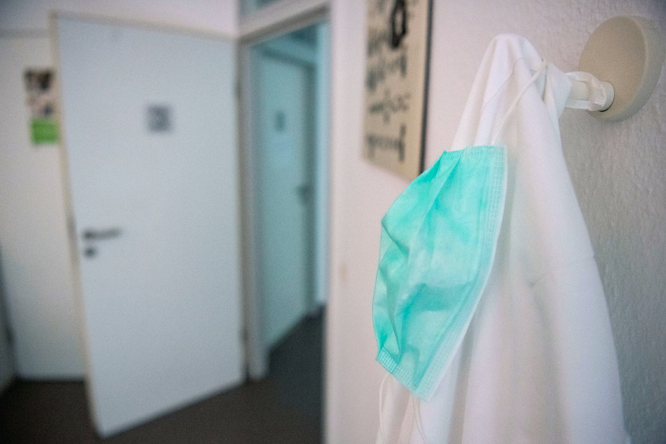Ein Mundschutz hängt in einer Hausarztpraxis in einem Behandlungszimmer über einem Arztkittel.