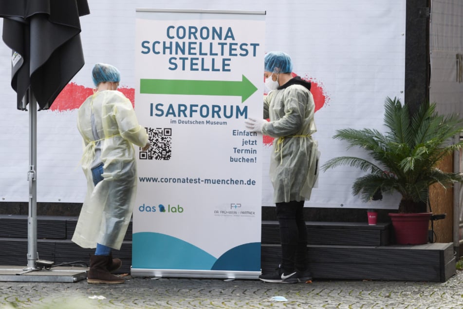 Vor der Corona-Teststelle am Deutschen Museum in München bauen zwei Mitarbeiterinnen in Schutzkleidung ein Rollup als Wegweiser auf.