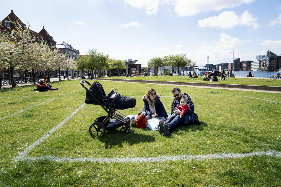 Eine Familie genießt die Sonne im Hafengebiet Islands Brygge. Markierte Flächen auf dem Grasland helfen den Menschen, Abstand voneinander zu halten, wobei etwa 40 Quadratmeter Fläche für maximal 10 Personen markiert sind.