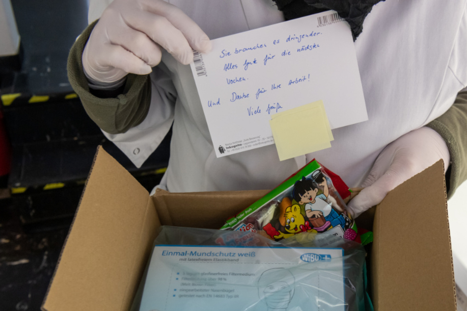 Eine Mitarbeiterin einer Hausarztpraxis im Münchner Stadtteil Haidhausen zeigt ein zugeschicktes Paket mit einer Packung Mundschutz, Gummibärchen und einer Karte mit der Aufschrit "Sie brauchen es dringender. Alles Gute für die nächsten Wochen. Und danke für ihre Arbeit!".
