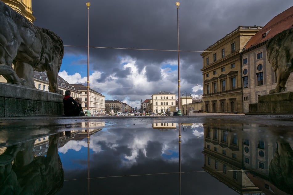 Wolken spiegeln sich in einer Pfütze in der Feldherrnhalle in Richtung Ludwigstraße, deren Bauten von der Sonne beschienen werden. (Archivbild)