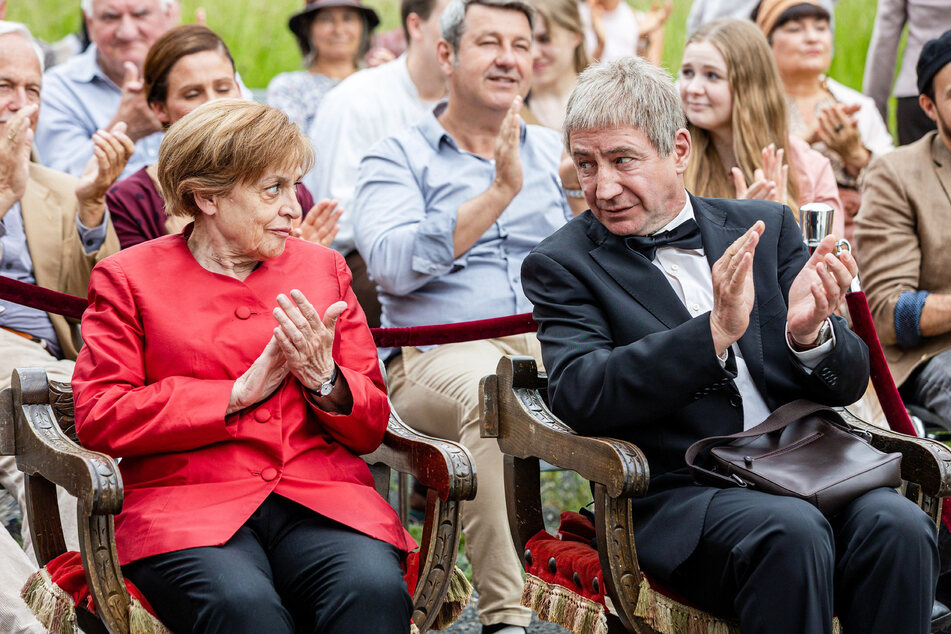 Merkel und ihr Ehemann Joachim Sauer (Thorsten Merten, 59) verfolgen gelangweilt das Schauspiel auf der Bühne.
