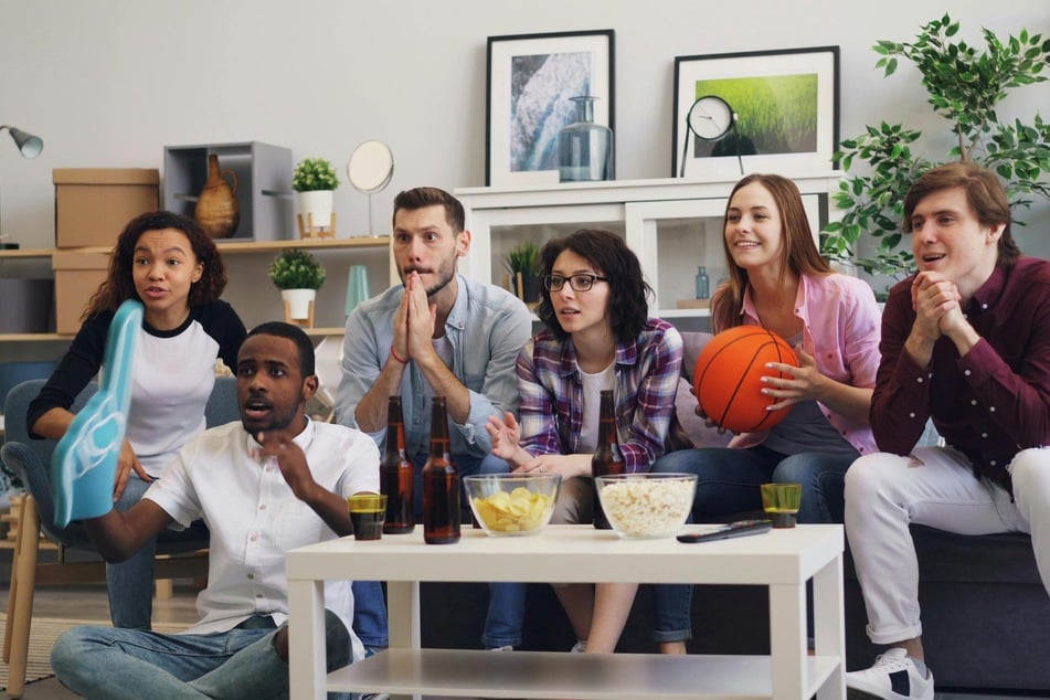 Sechs junge Erwachsene sitzen auf einem Sofa und auf dem Boden vor dem TV. Eine Person hält einen Foam-Finger hoch, eine andere einen Basketball.