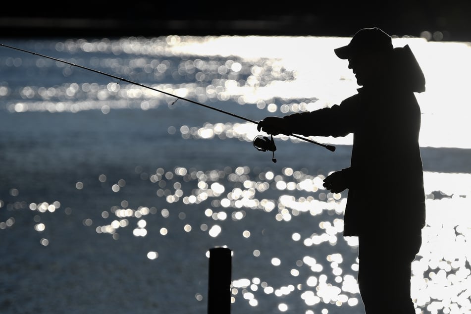 Ein Angler steht am späten Nachmittag am Ufer eines Flusses im Gegenlicht (Symbolbild).
