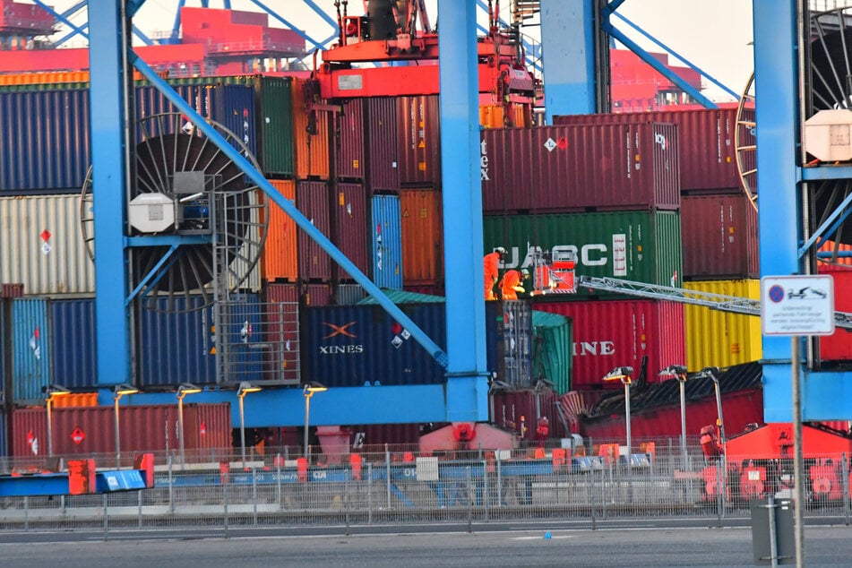 Feuerwehrleute in Schutzanzügen untersuchen die beschädigten Container im Hafen.