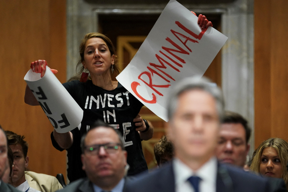 A pro-Palestine protester with a sign reading "Criminal" is removed by police during Secretary of State Antony Blinken's testimony before the Senate Foreign Relations Committee.