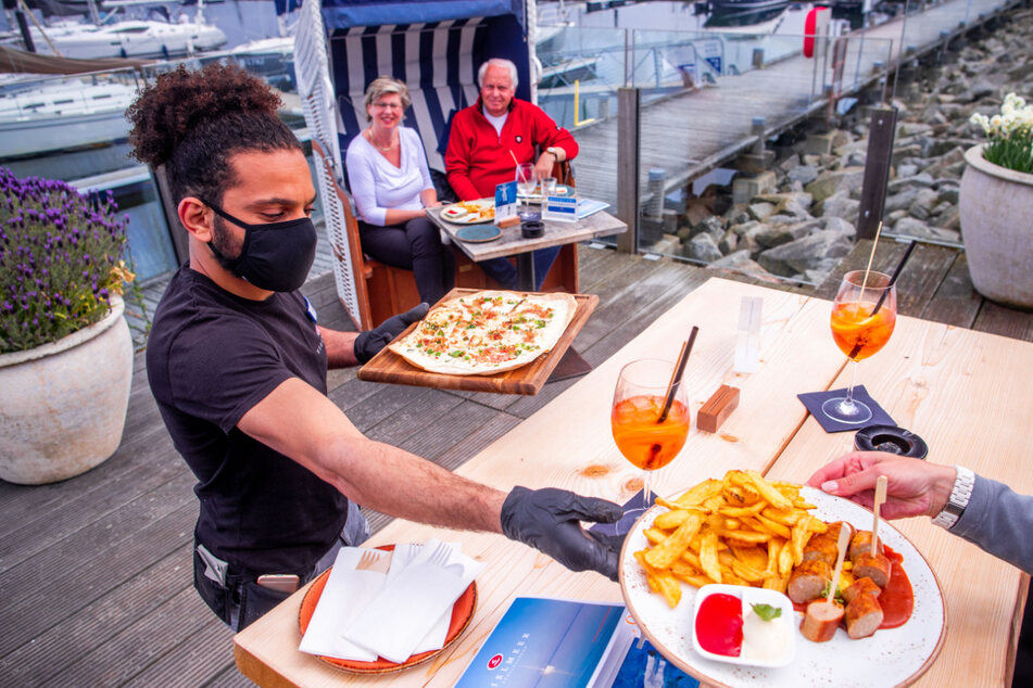 Eine Kellner serviert mit Mundschutz und Handschuhen seinen Gästen das Essen.