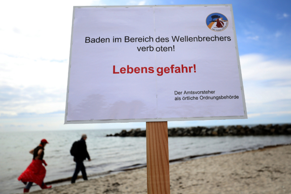 Spaziergänger am Ostseestrand Ahrenshoop. Im Vordergrund steht ein Warnschild. (Symbolbild)
