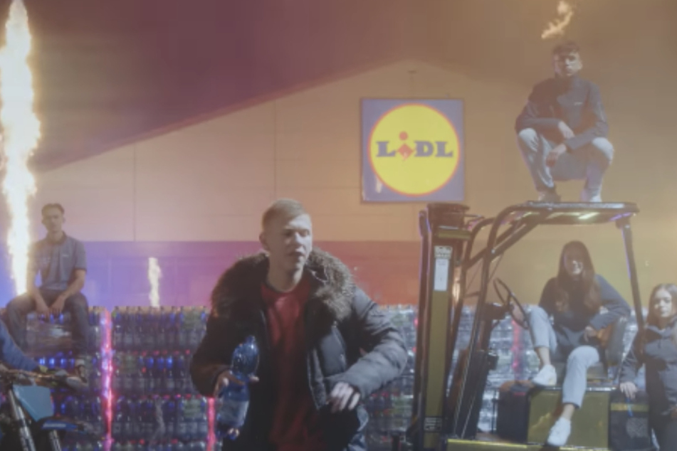Screenshot aus dem Video: Im Hintergrund steigen auf dem Lidl-Parkplatz Feuerfontänen in die Luft, während Tillmann seinen Song zum Besten gibt.