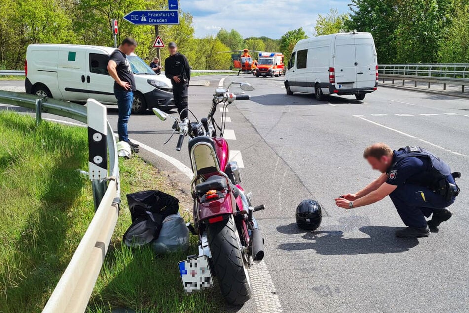 Ein Polizist dokumentiert die Schäden an einem am Boden liegenden Motorradhelm. Der in den Unfall verwickelte Kleintransporter ist rechts im Bild zu sehen. Im Hintergrund ist einer der beiden Rettungshubschrauber auszumachen.