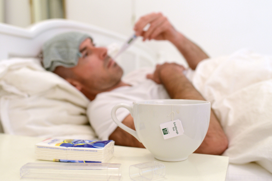 Ein kranker Mann liegt im Bett und misst Fieber. In Sachsen hat die Grippe schon mehr als 14.000 Menschen erwischt.