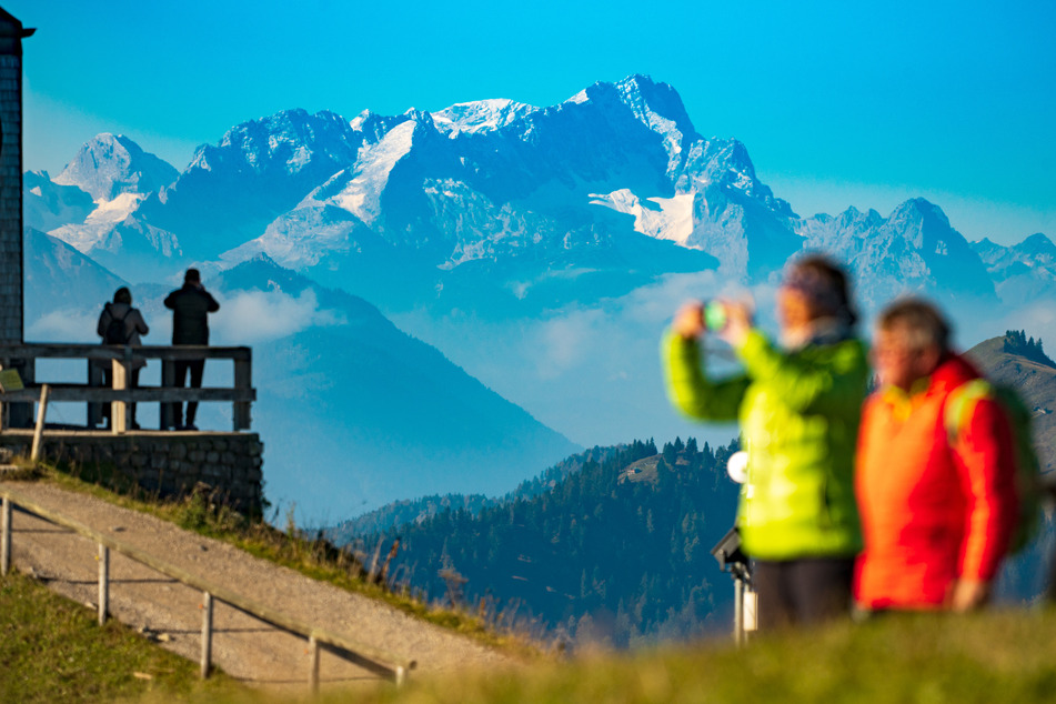 Besucher stehen in den Morgenstunden auf dem Wallberg vor der imposanten Kulisse der Zugspitze, die im Hintergrund in den Himmel ragt.