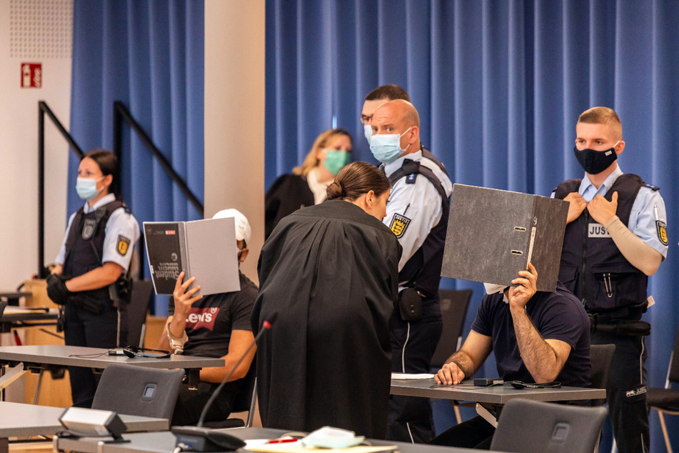 Zwei Angeklagte halten Mappen als Sichtschutz vor ihre Gesichter, während eine Verteidigerin sich mit dem rechten Angeklagten bespricht.