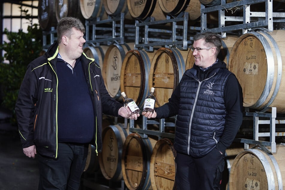Sippel (r.) und Lenhardt stehen vor Whiskyfässern und halten Flaschen mit zum Whisky gebrannten Bier in den Händen.