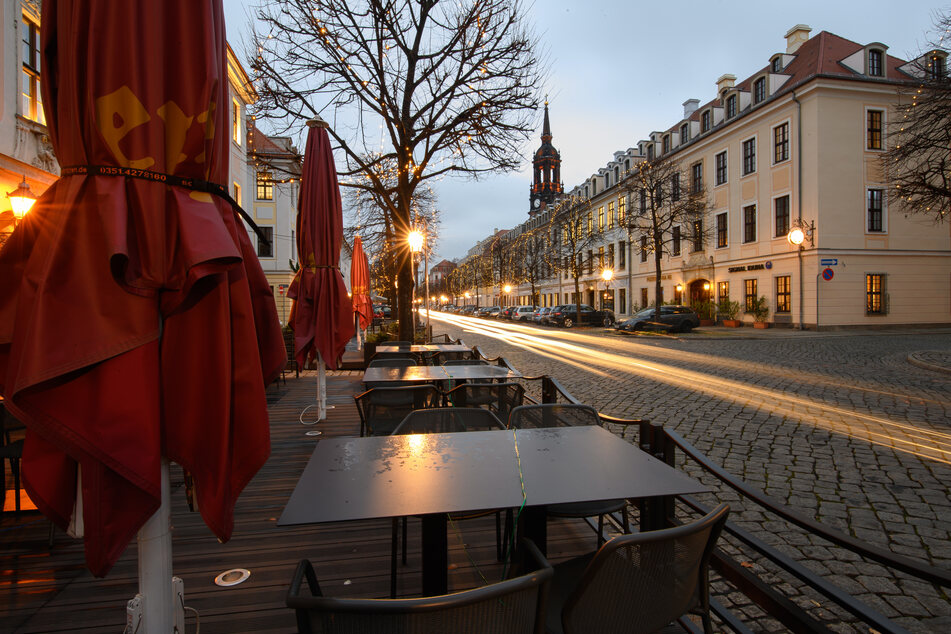 Sachsen, Dresden: Leere Tische und geschlossene Schirme stehen vor einem Restaurant auf während auf der Königstraße die Scheinwerfer vorbeifahrender Autos Lichtspuren ziehen.