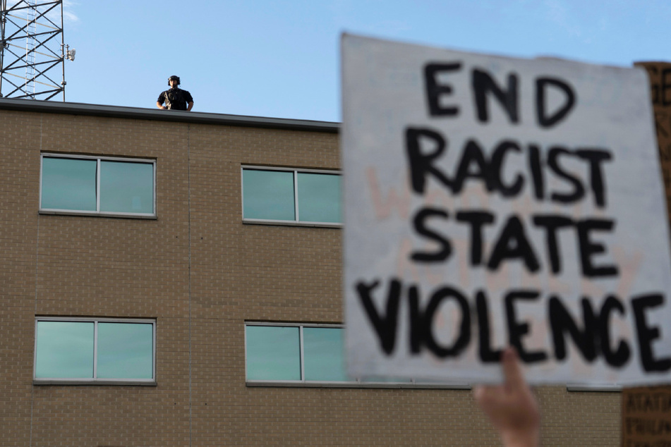 Ein Polizeibeamter steht am Samstag auf dem Dach einer Polizeidienststelle in Aurora, während bei einer Demonstration gegen den Tod von Elijah McClain ein Plakat mit der Aufschrift "End Racist State Violence" ("Beendet rassistische staatliche Gewalt") in die Höhe gehalten wird. McClain starb Ende August 2019, nachdem er auf dem Weg zu seiner Wohnung von drei Polizisten von Aurora angehalten wurde.