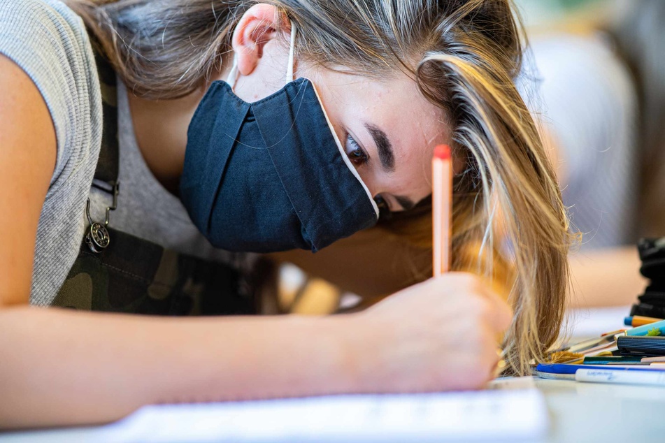 Eine Schülerin sitzt mit Mund-Nasen-Bedeckung am Tisch und schreibt.