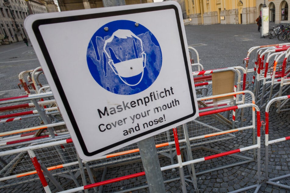 Zusammengestellte Absperrgitter stehen am Münchner Odeonsplatz hinter einem Schild mit der Aufschrift "Maskenpflicht . Cover your mouth an nose!".