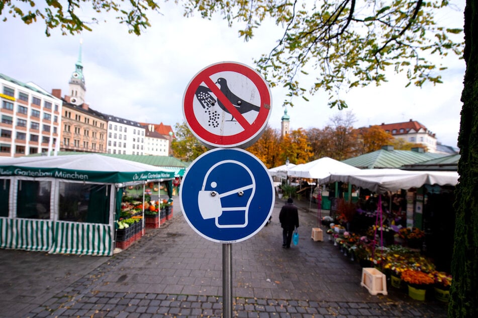 Eine symbolische Darstellung der Maskenpflicht ist auf einem Hinweisschild auf dem Viktualienmarkt in der Innenstadt zu sehen.